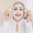 Close up beauty portrait of smiling pretty senior gray haired woman with facial mud mask and cucumber slices in her hands, posing on light home interior background. Skin cosmetics and spa concept.